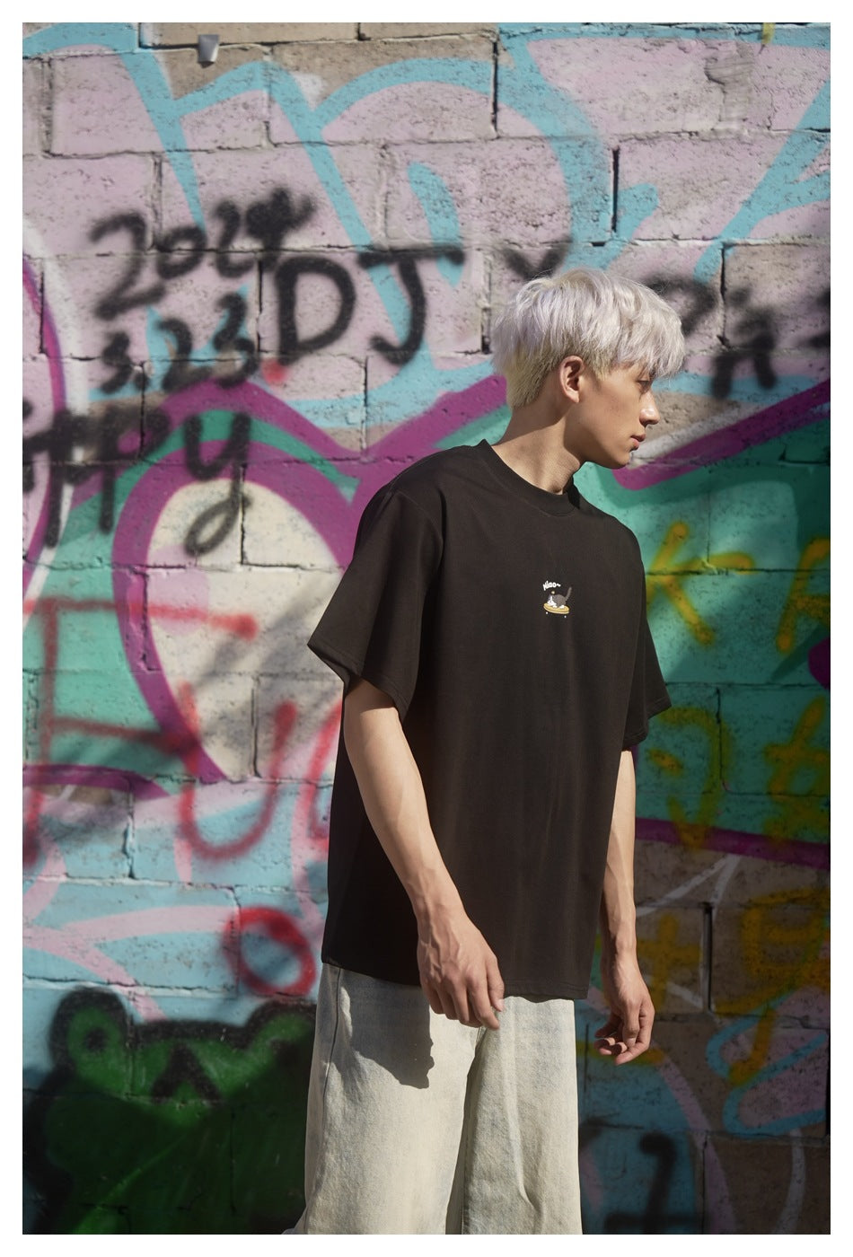 Model wearing a black oversized T-shirt with a small cat-on-skateboard graphic, shown from the side against a graffiti wall.