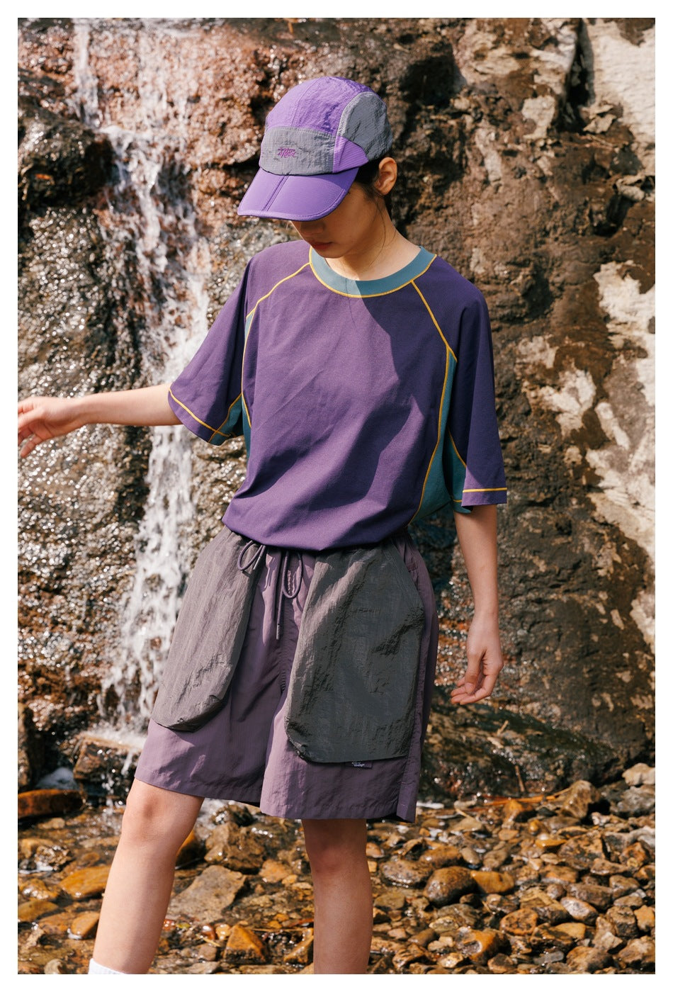 Close view of a model wearing a purple shirt with teal panels and yellow piping, with matching shorts beside a rocky stream.