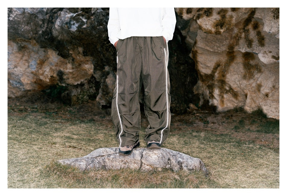 Front view of a model wearing loose taupe-gray pants with bright reflective side piping, outdoors.