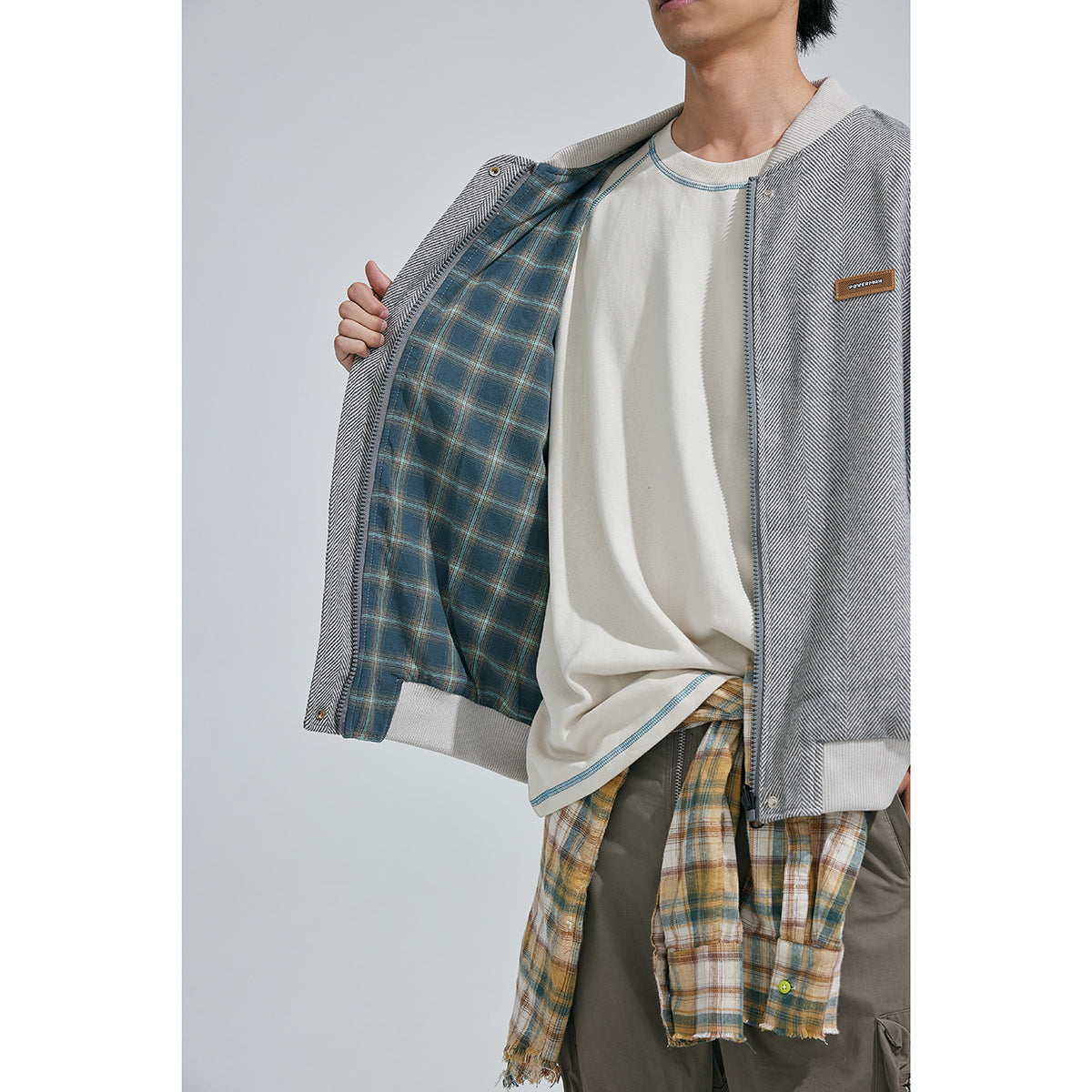 Model holds open a gray herringbone bomber jacket, showing its blue-and-tan plaid lining.