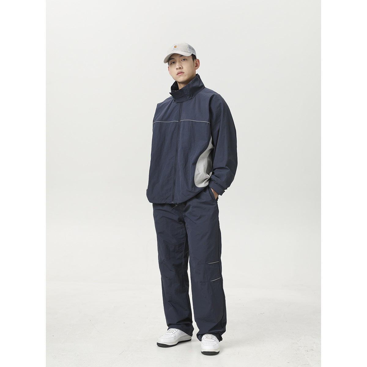 Model wearing a navy zip jacket with reflective piping and matching loose-fit pants, white sneakers, and a light cap.
