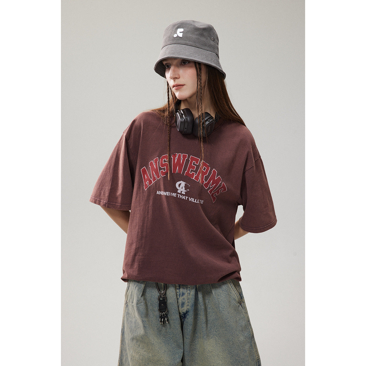 Model wearing an oversized burgundy graphic T-shirt with loose blue jeans and a gray bucket hat.