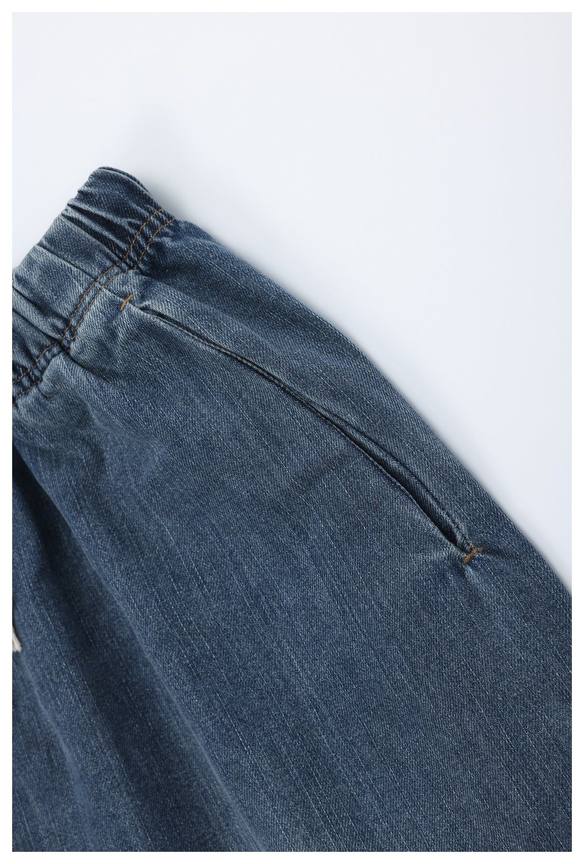 Close-up of blue denim trousers showing a pocket opening, seam, and brown stitching.