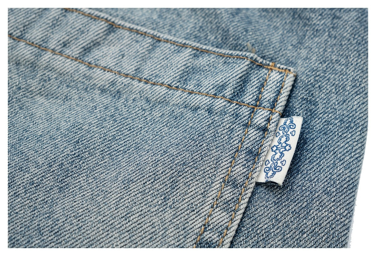 Close-up of a rear jeans pocket seam with tan stitching and a small white fabric tab bearing a blue ornamental motif.