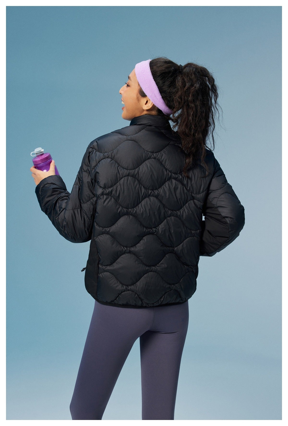 Rear view of a woman wearing a black wave-quilted jacket and purple leggings, holding a purple bottle.