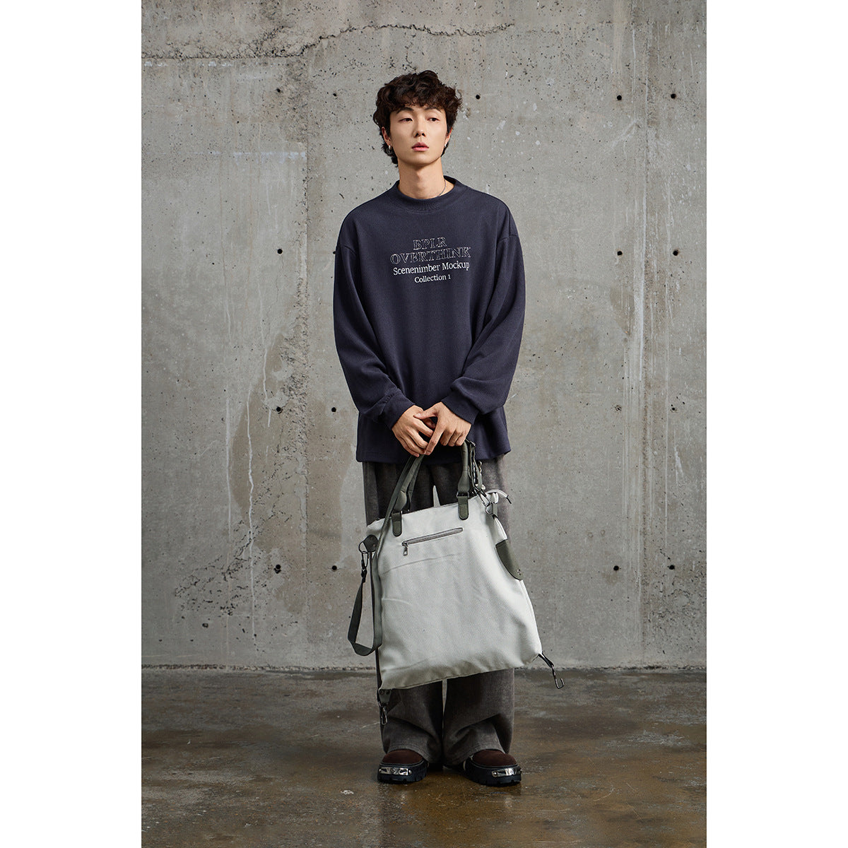 Model wearing an oversized navy crewneck with white chest lettering, wide gray trousers, and carrying a large light tote.