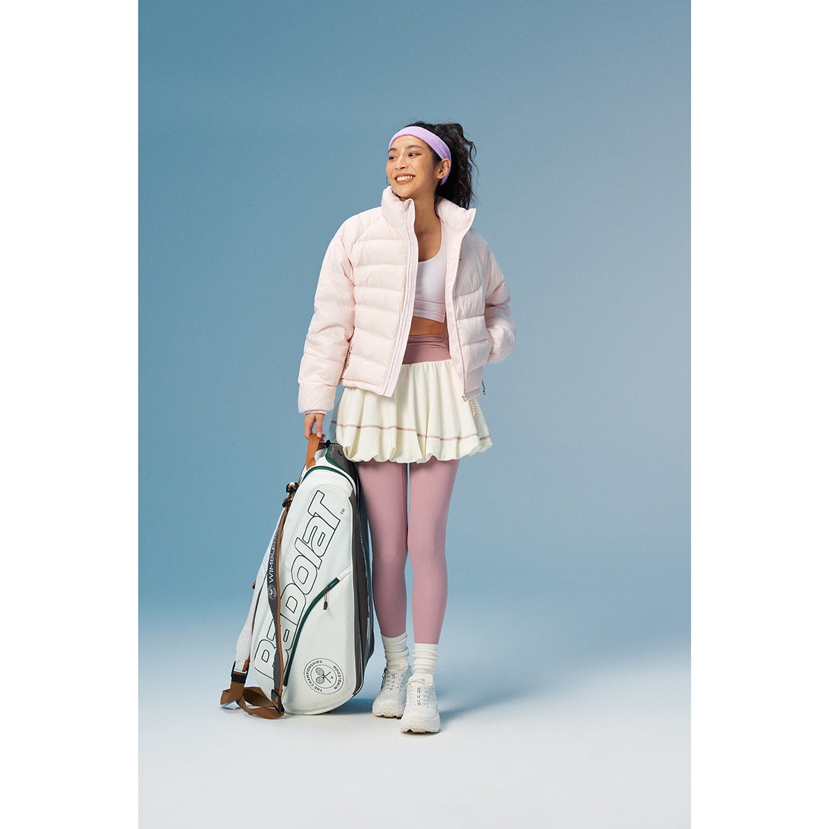 Model wearing a pale pink puffer jacket with a white tennis skirt and mauve leggings, holding a racquet bag.