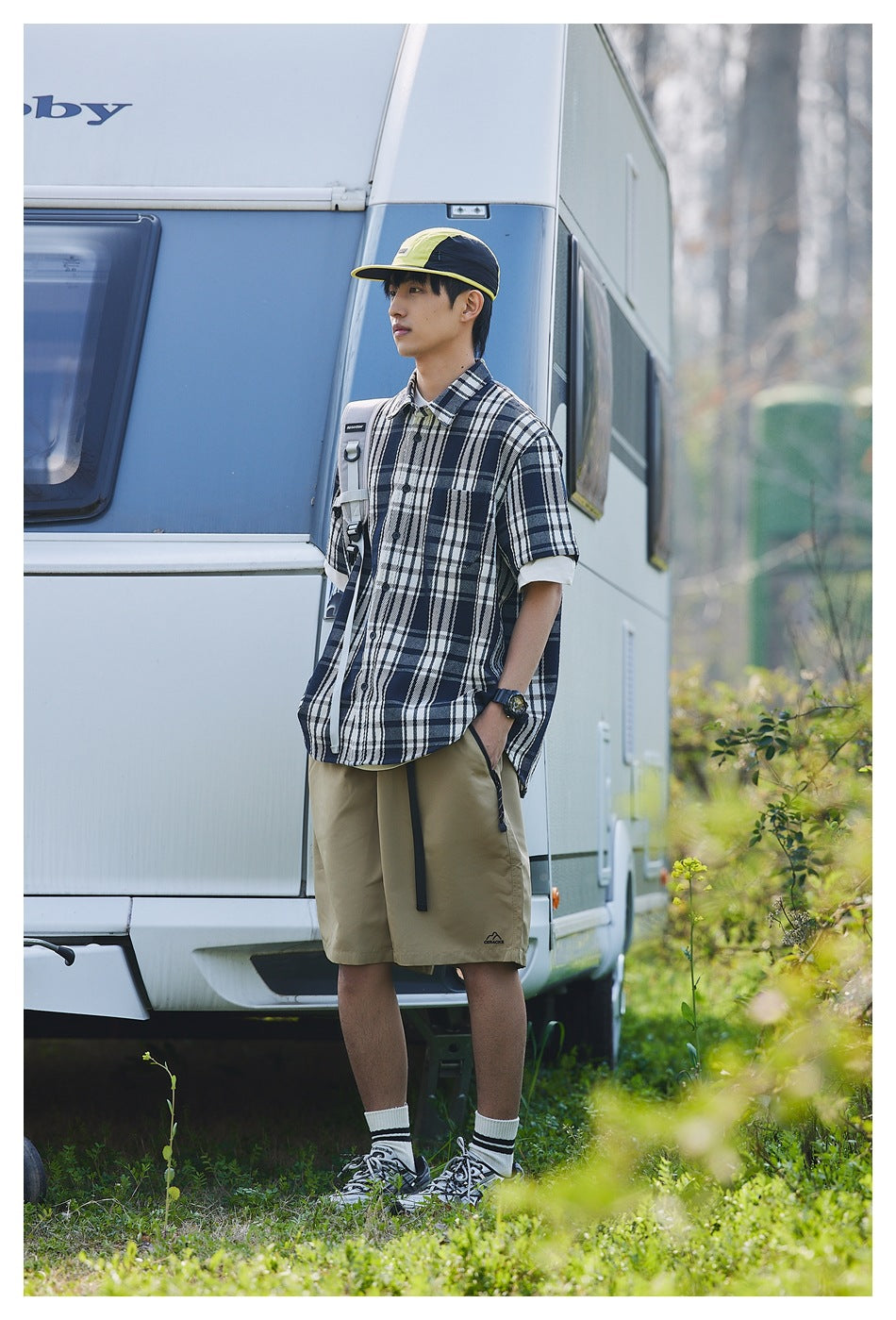 Man in navy plaid short-sleeve shirt and khaki shorts standing beside a camper trailer outdoors