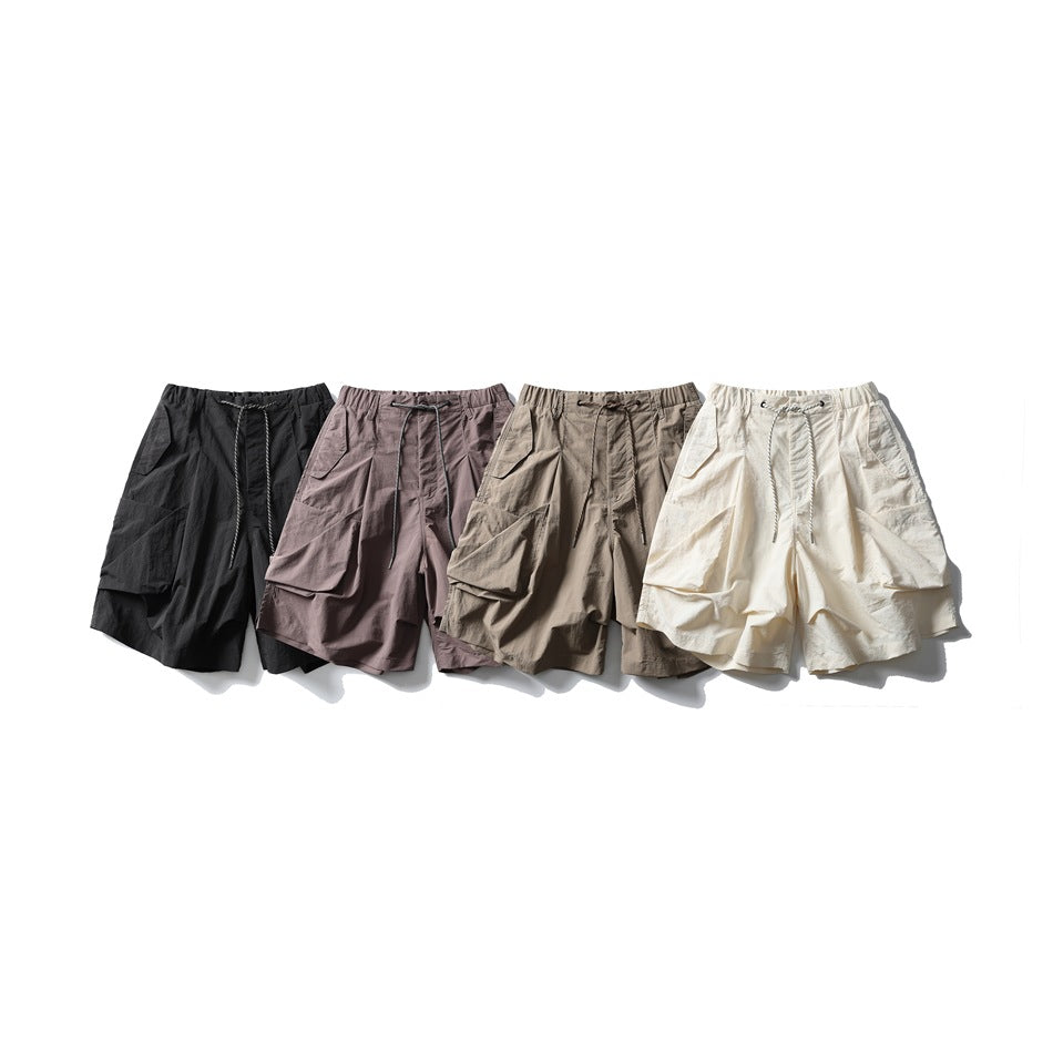 Four pairs of wide drawstring cargo shorts shown together in black, mauve-gray, taupe, and cream.