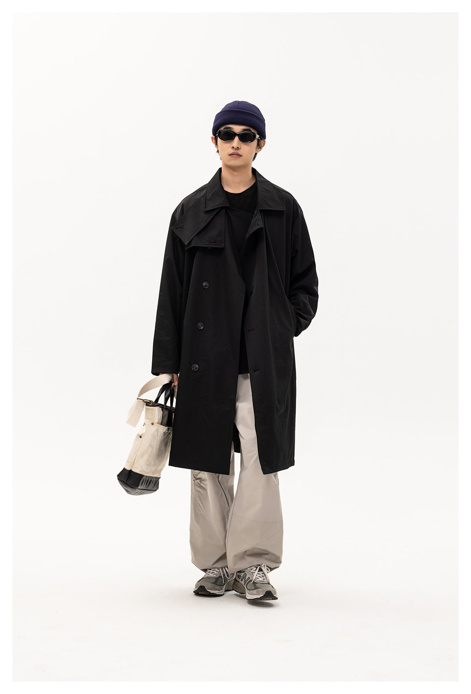 Model wearing a black loose long trench coat with a chest flap, light gray wide pants and a navy beanie, holding a tote
