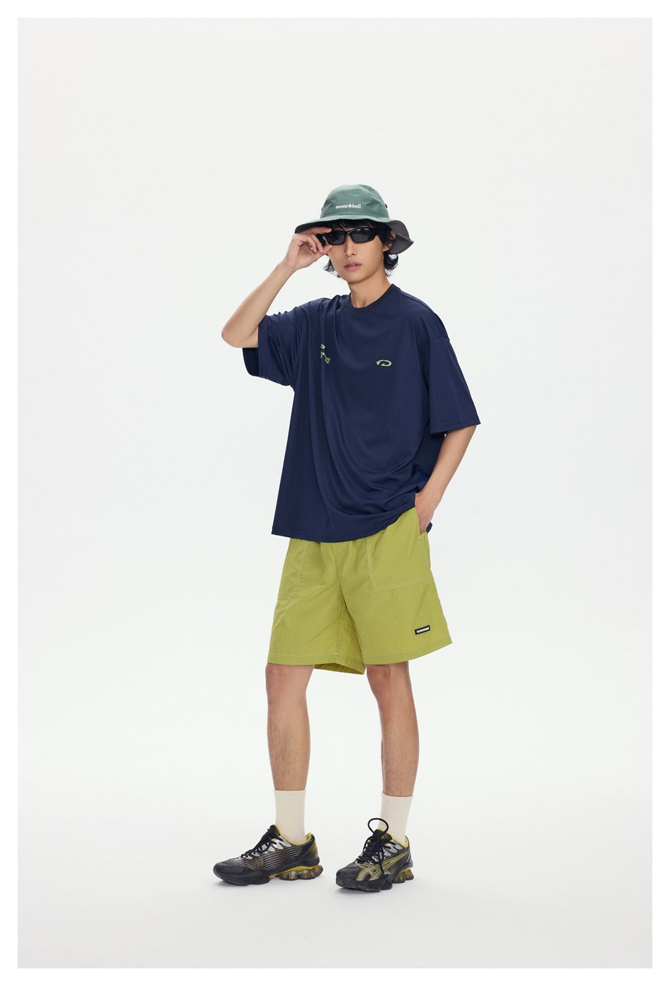 Full-length model wearing a navy graphic T-shirt and lime shorts with a green bucket hat and dark sneakers.