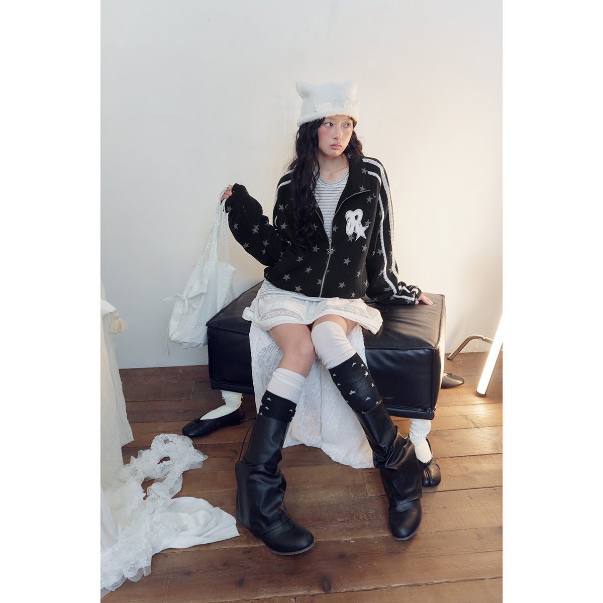 Seated model wearing a black star-print zip jacket with white sleeve stripes and a white chest patch.