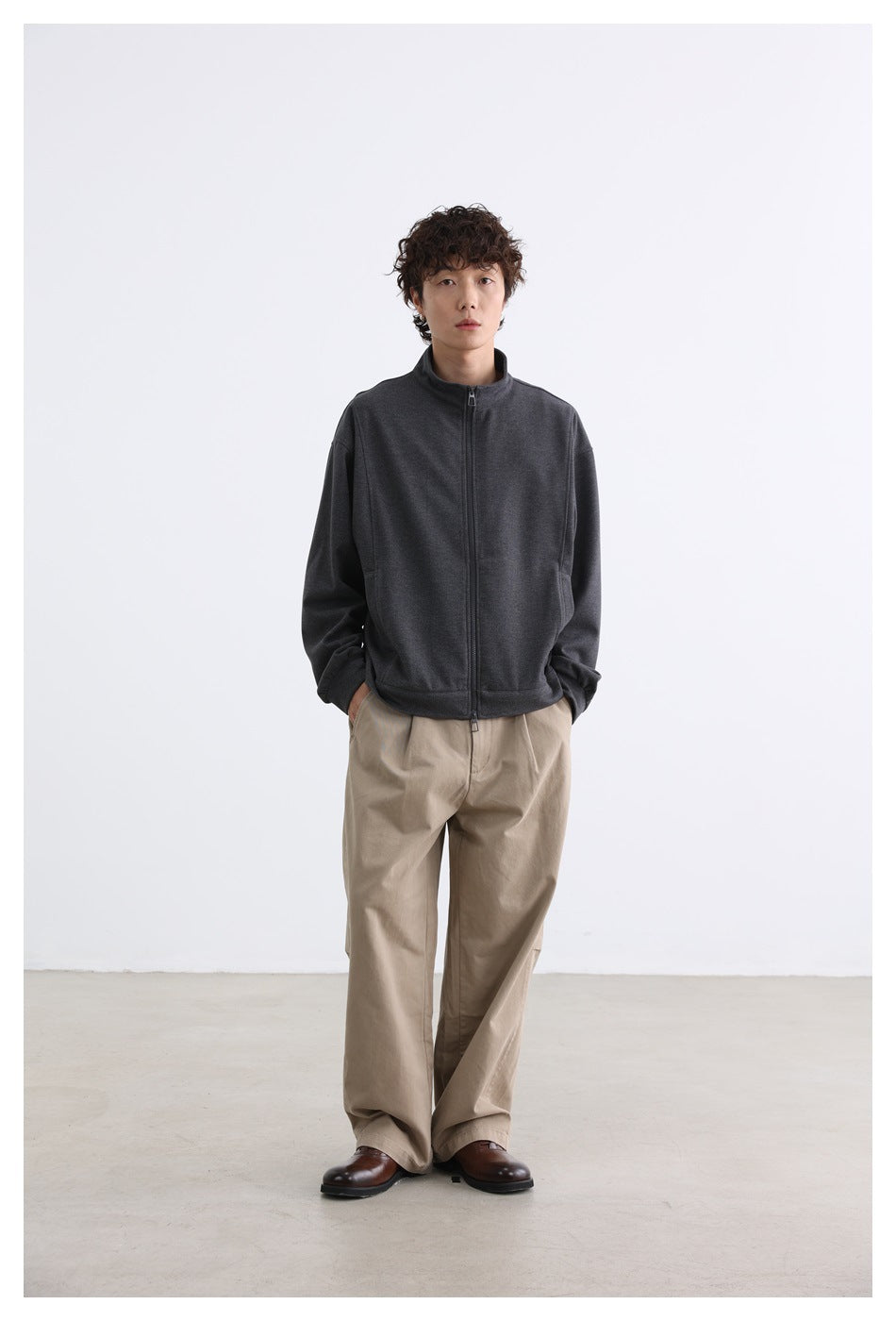 Full-length view of a model wearing a charcoal stand-collar zip jacket and beige trousers.