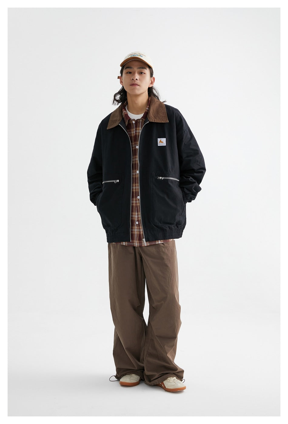 Model wearing the black zip jacket open over a plaid shirt with brown wide-leg pants and a cap, standing front view