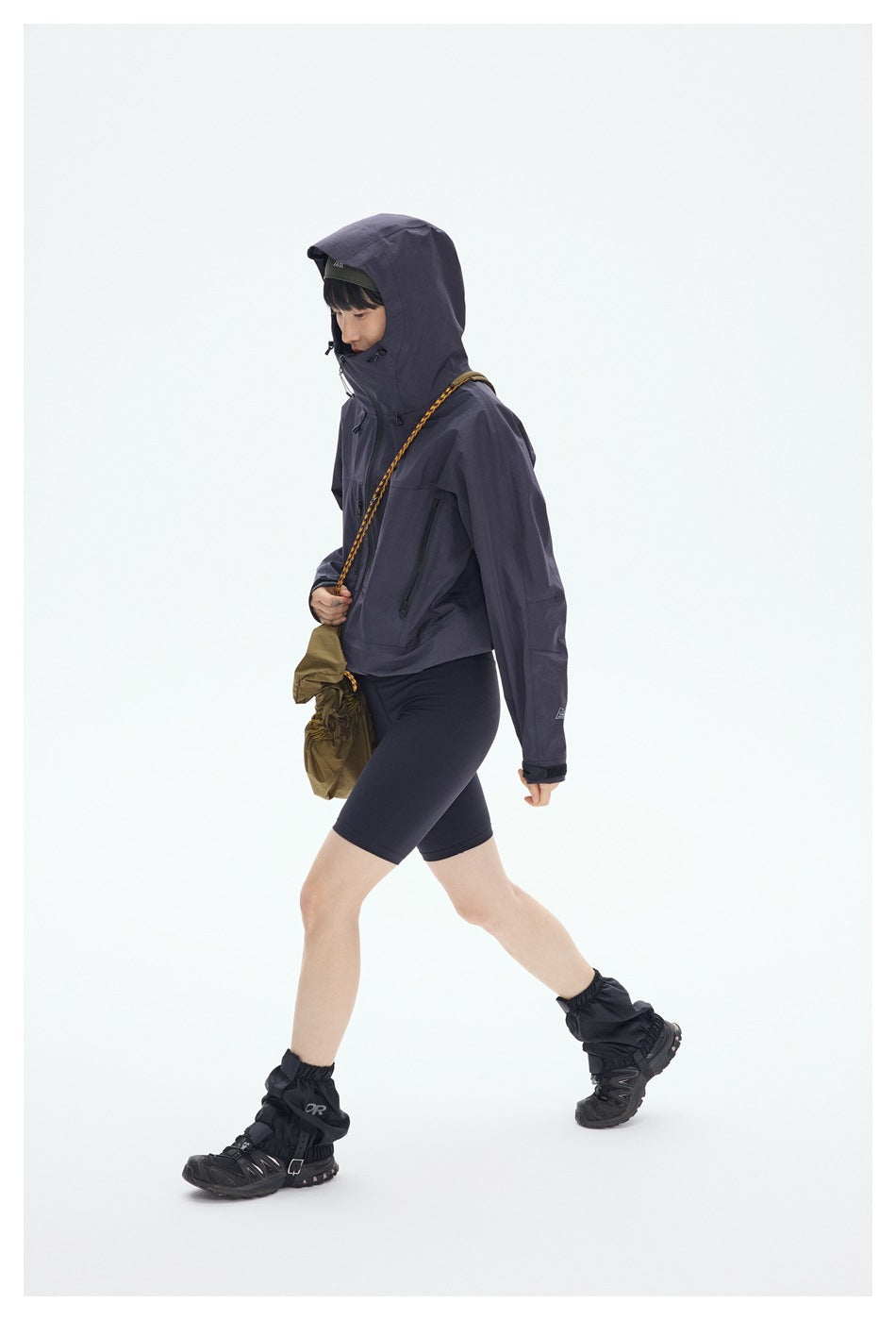 Model walks in profile wearing the dark plum jacket with its hood up, black shorts and hiking gaiters.