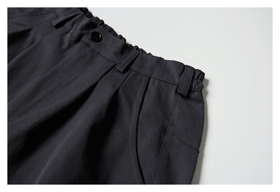 Close-up of charcoal trousers showing the buttoned waistband, belt loops, front pleat, and curved side pocket.