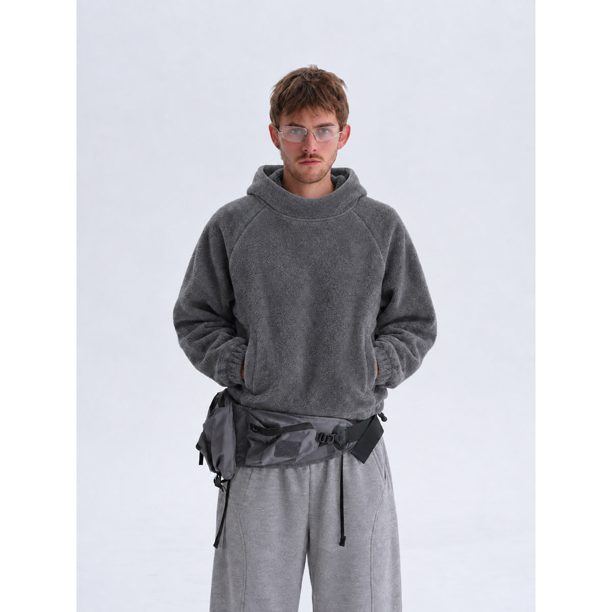 Model in a dark gray textured hooded sweatshirt and matching loose gray pants, with a dark utility bag at the waist.