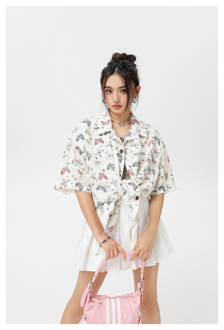 Front view of a model in a cream butterfly-print short-sleeve shirt tied at the waist with a white pleated skirt.