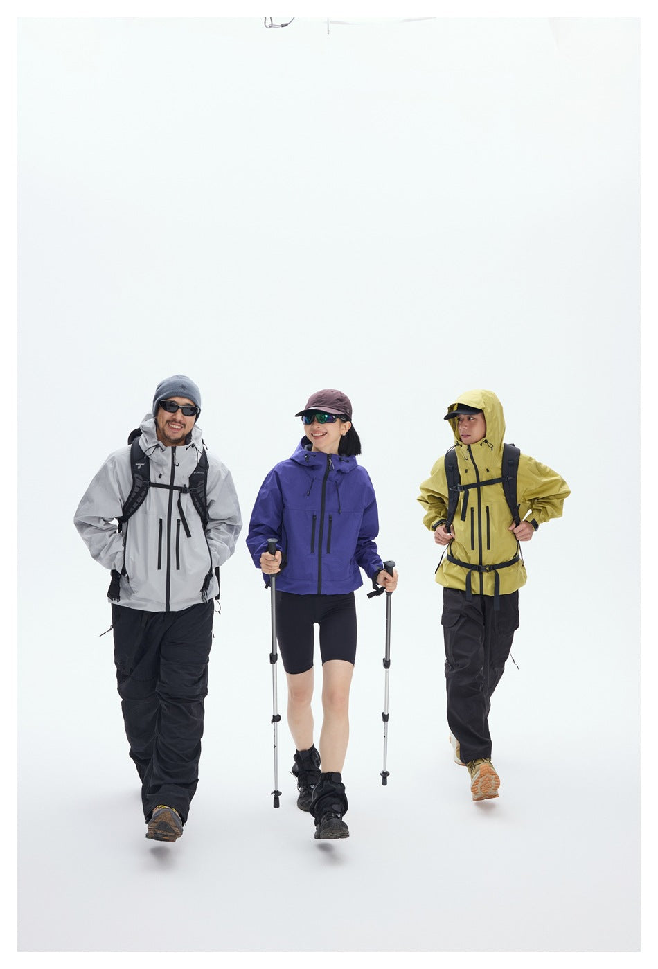 Three hikers model gray, purple, and yellow hooded jackets with black pants and trekking poles against a white backdrop.