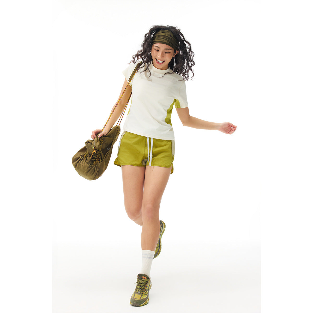 Model wears a white T-shirt with lime side panels and olive-yellow shorts, holding a matching olive bag.