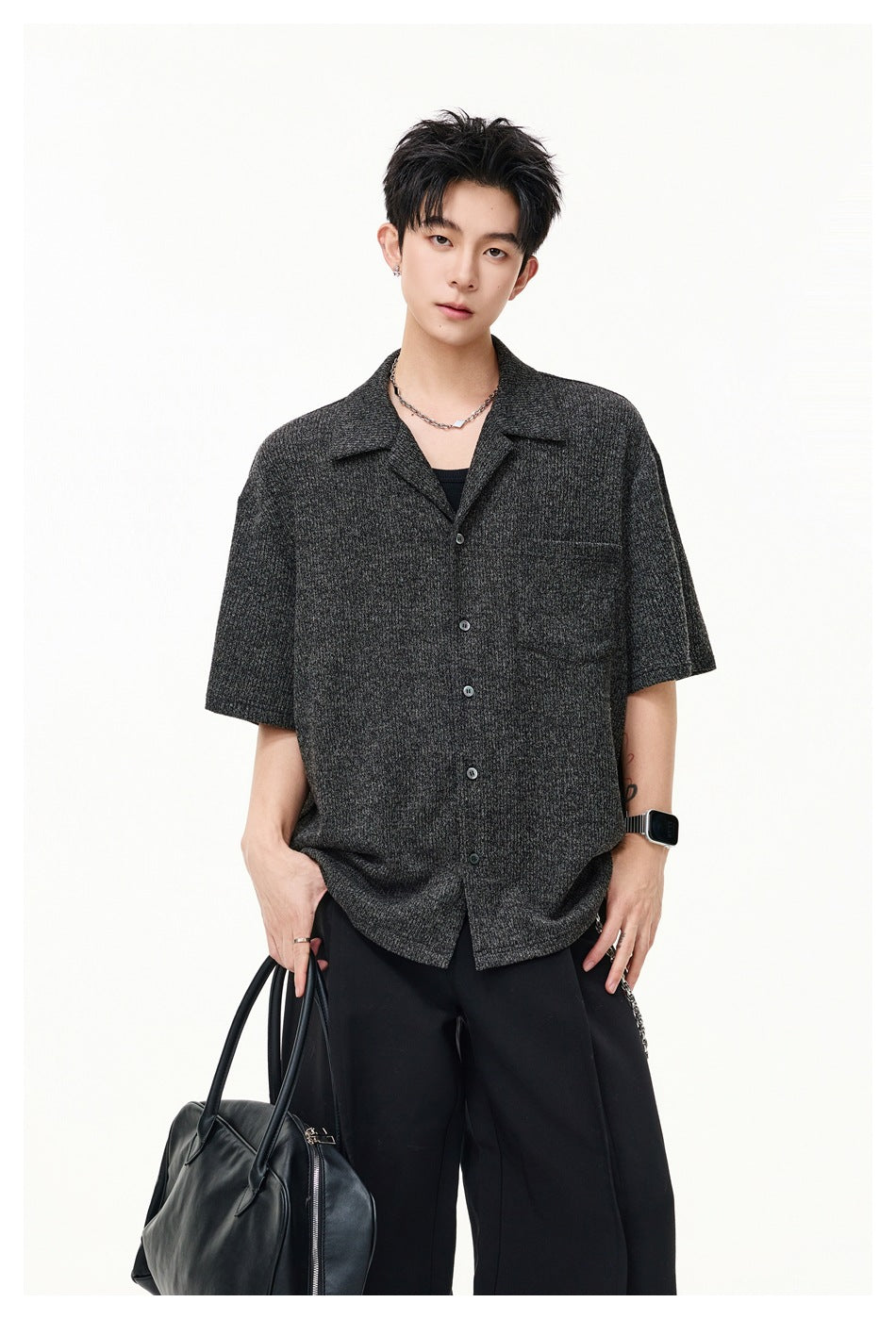 Man wearing a gray textured short-sleeve camp-collar shirt with black trousers, holding a black handbag