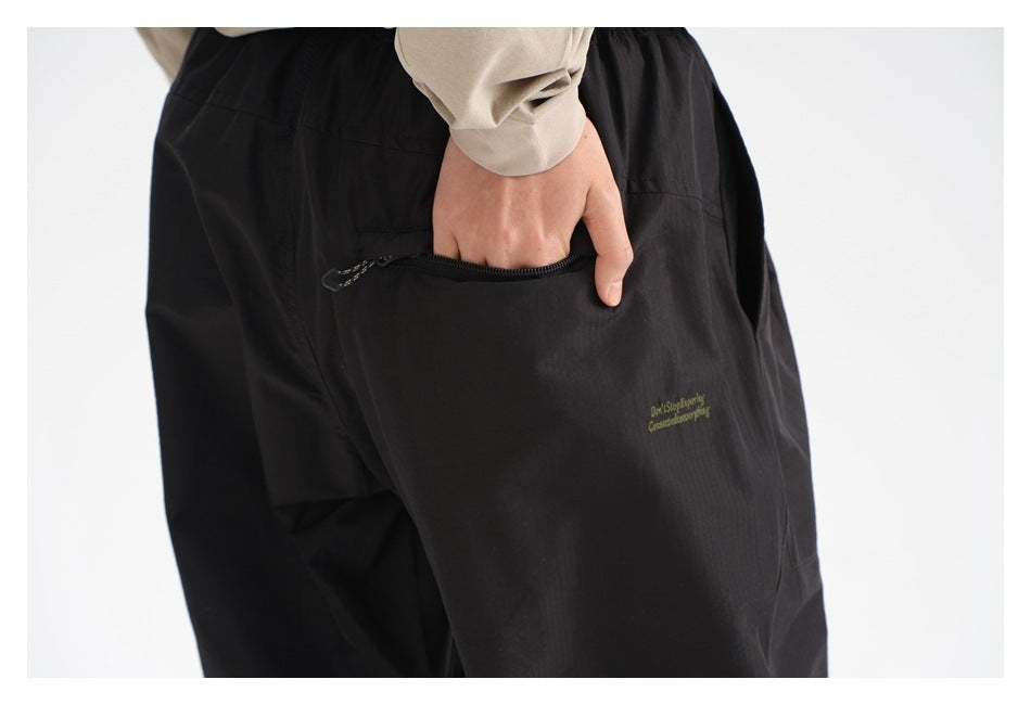 Close-up of black pants showing a zippered pocket, small embroidered text, and a hand in the pocket.
