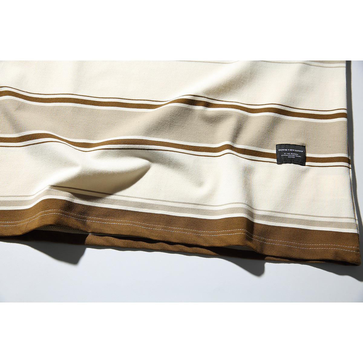 Close-up of the striped T-shirt's lower hem, showing cream, taupe, and brown bands and a small black sewn label.