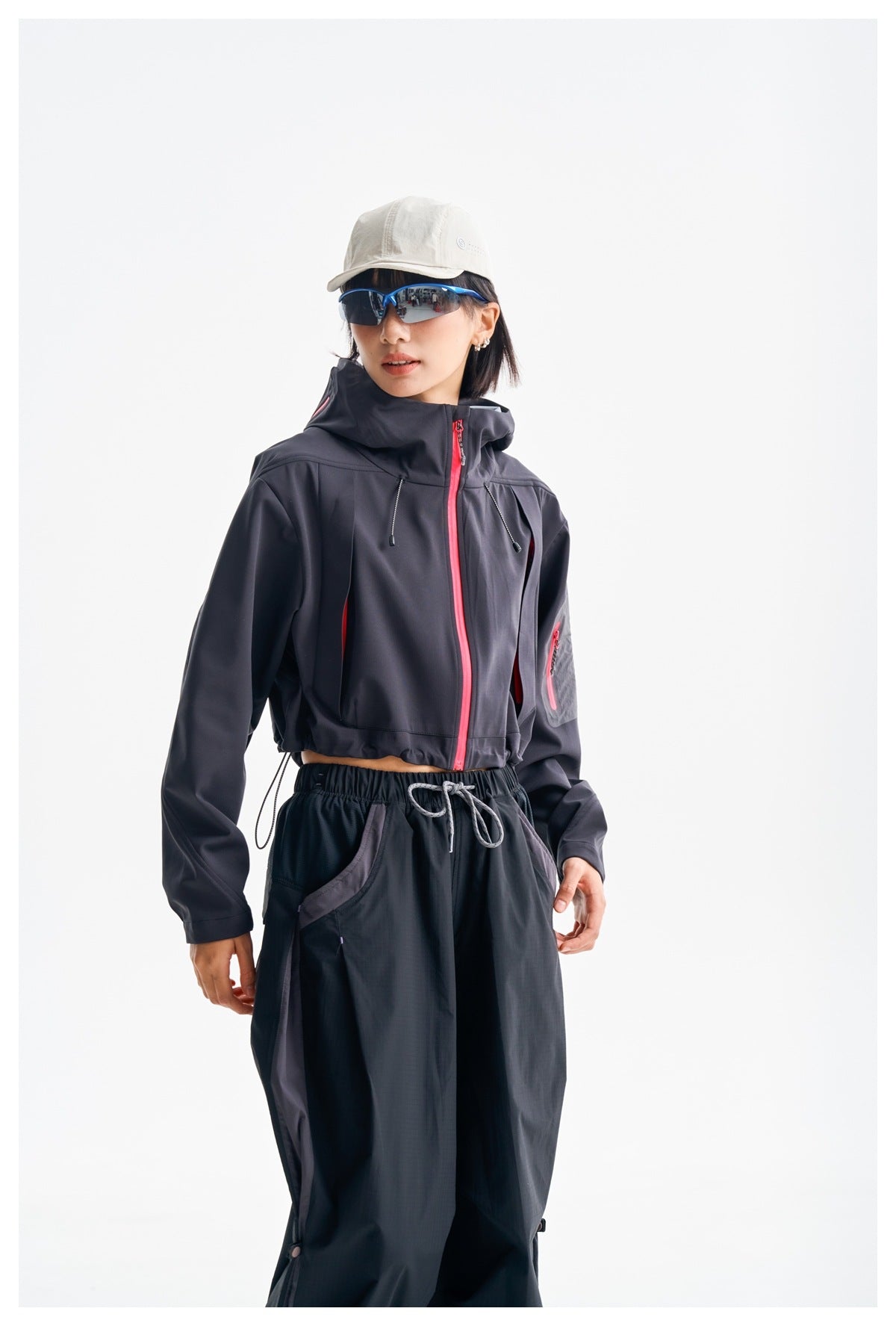 Model wearing a dark cropped hooded jacket with bright pink zippers and loose black trousers.