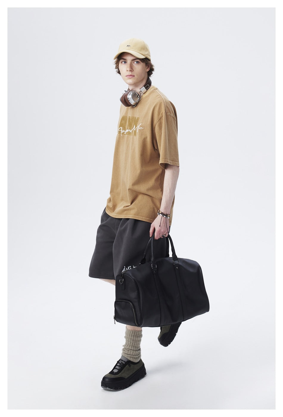Model in a tan graphic T-shirt, black shorts, beige cap and sneakers, carrying a large black duffel bag.