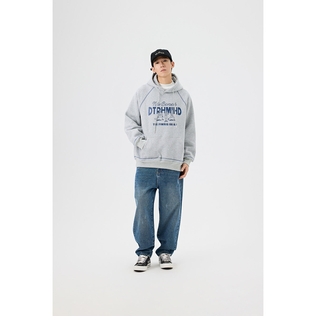 Full-length model wearing a light gray graphic hoodie, loose blue jeans, and black-and-white sneakers.