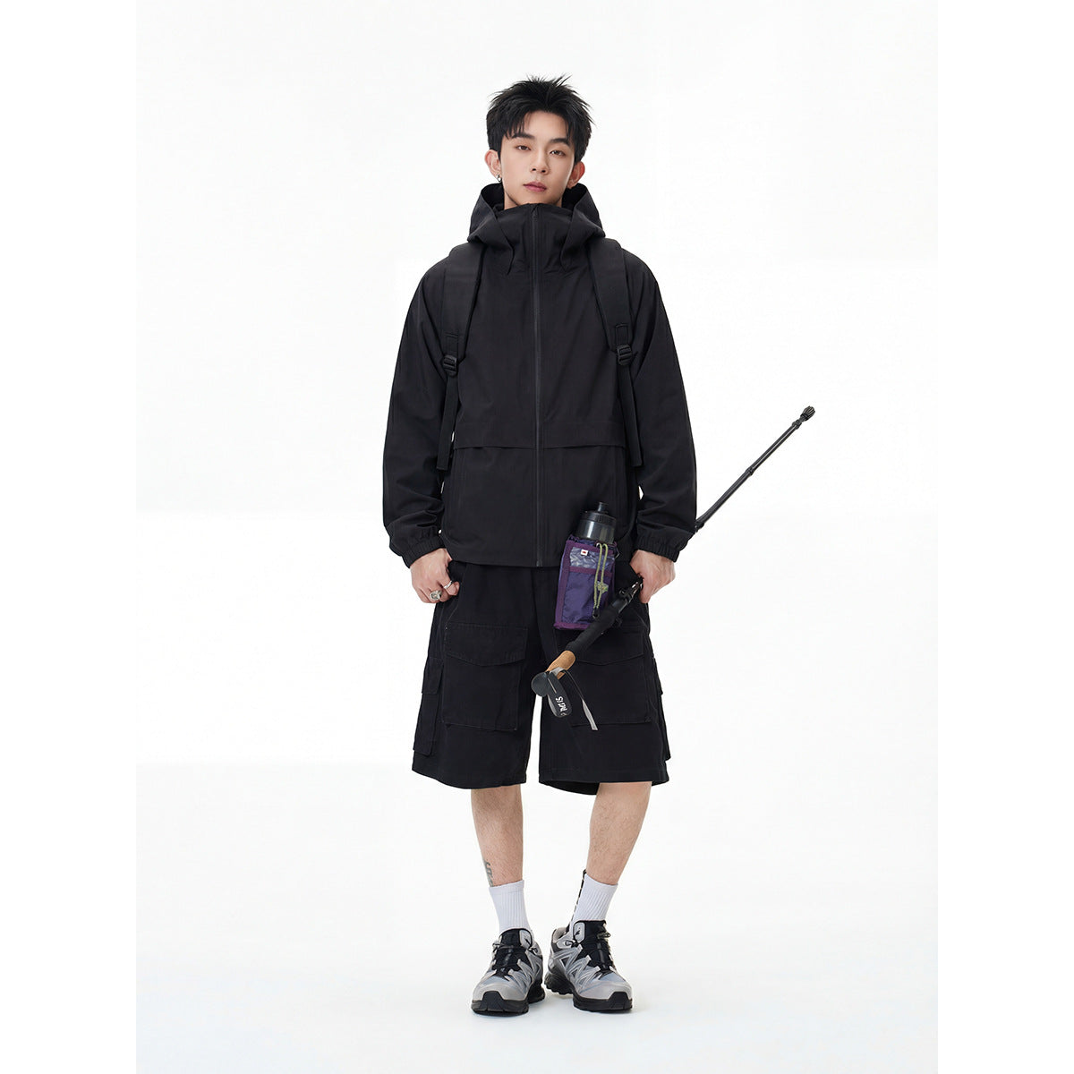 Model wearing a black hooded zip jacket and cargo shorts with a backpack and fishing gear.