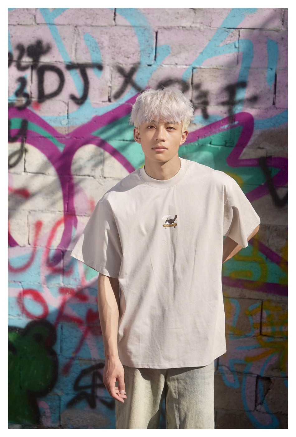 Front view of a model wearing a light beige oversized T-shirt with a small cat-on-skateboard chest graphic.
