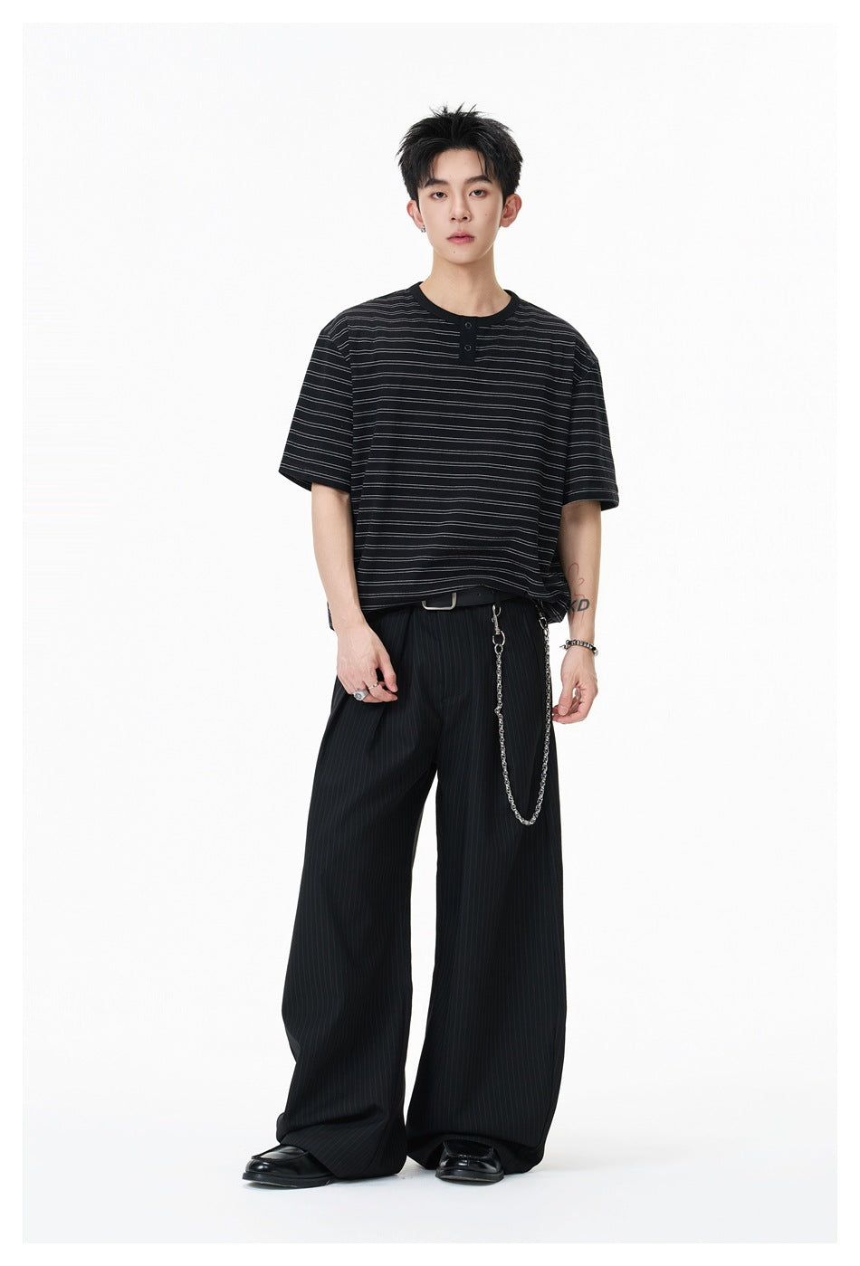 Model wearing a black short-sleeve shirt with thin horizontal stripes and a two-button placket, with wide pinstripe trousers.