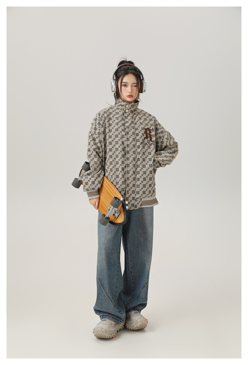 Full-length model wearing a beige-and-charcoal checker jacket with wide-leg jeans and holding a skateboard.