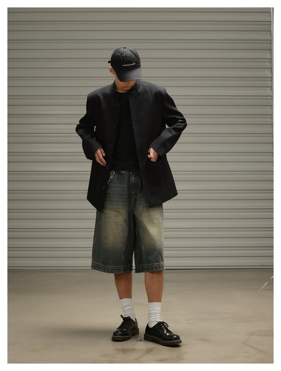Model in a black band-collar coat and oversized faded denim shorts, looking down while adjusting the coat.