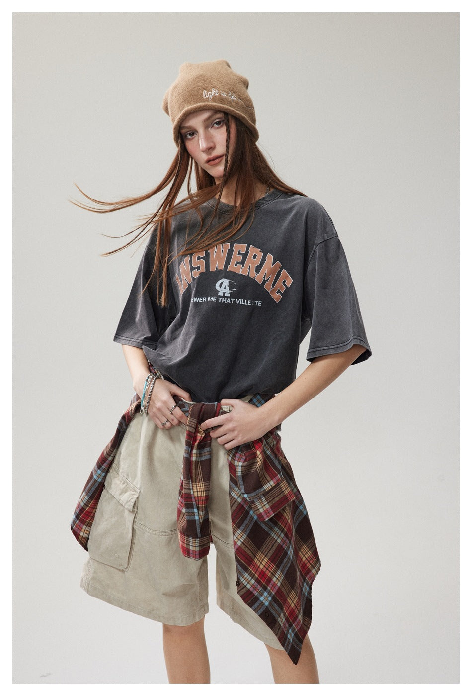 Model wearing an oversized charcoal graphic T-shirt with beige cargo shorts and a red plaid shirt tied at the waist.