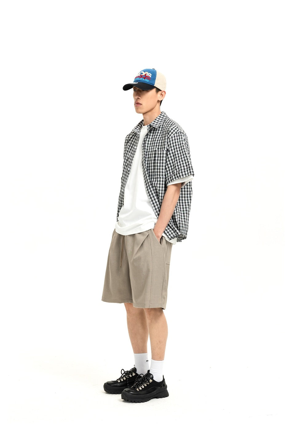 Model wearing khaki drawstring shorts with a plaid short-sleeve shirt, white tee, cap and black shoes, side view