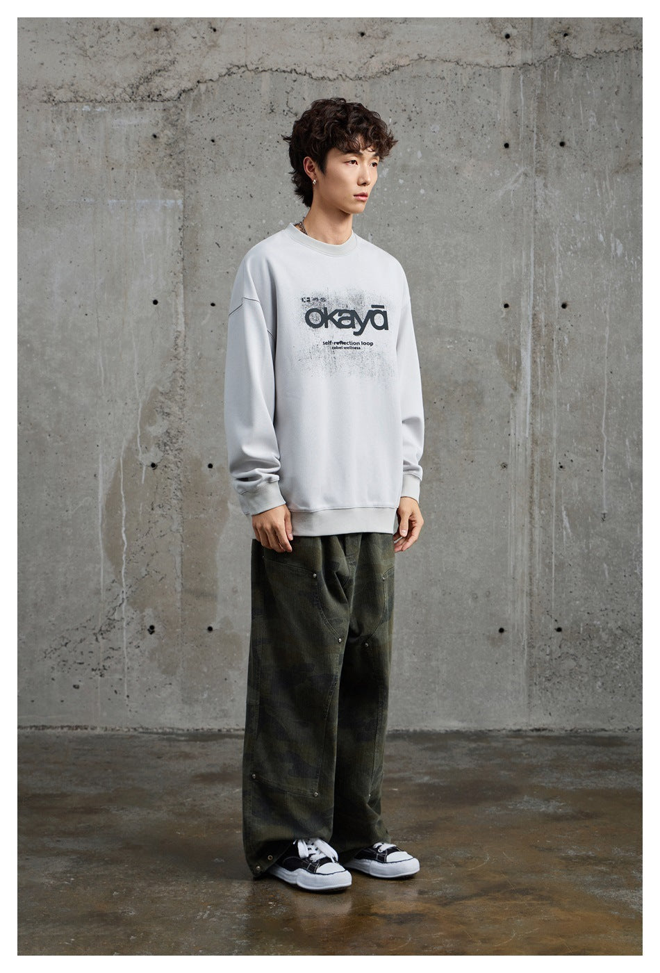 Side-front view of a model in a light gray graphic sweatshirt and loose dark camouflage pants.