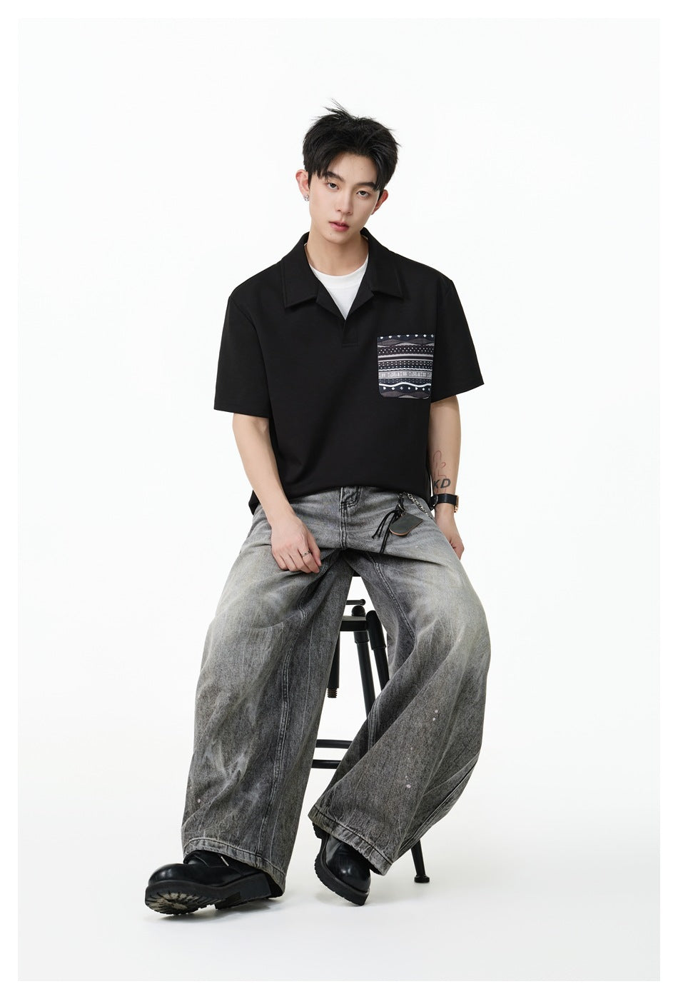 Model seated on a stool in a black short-sleeve polo-style top with patterned chest pocket and faded gray wide-leg jeans