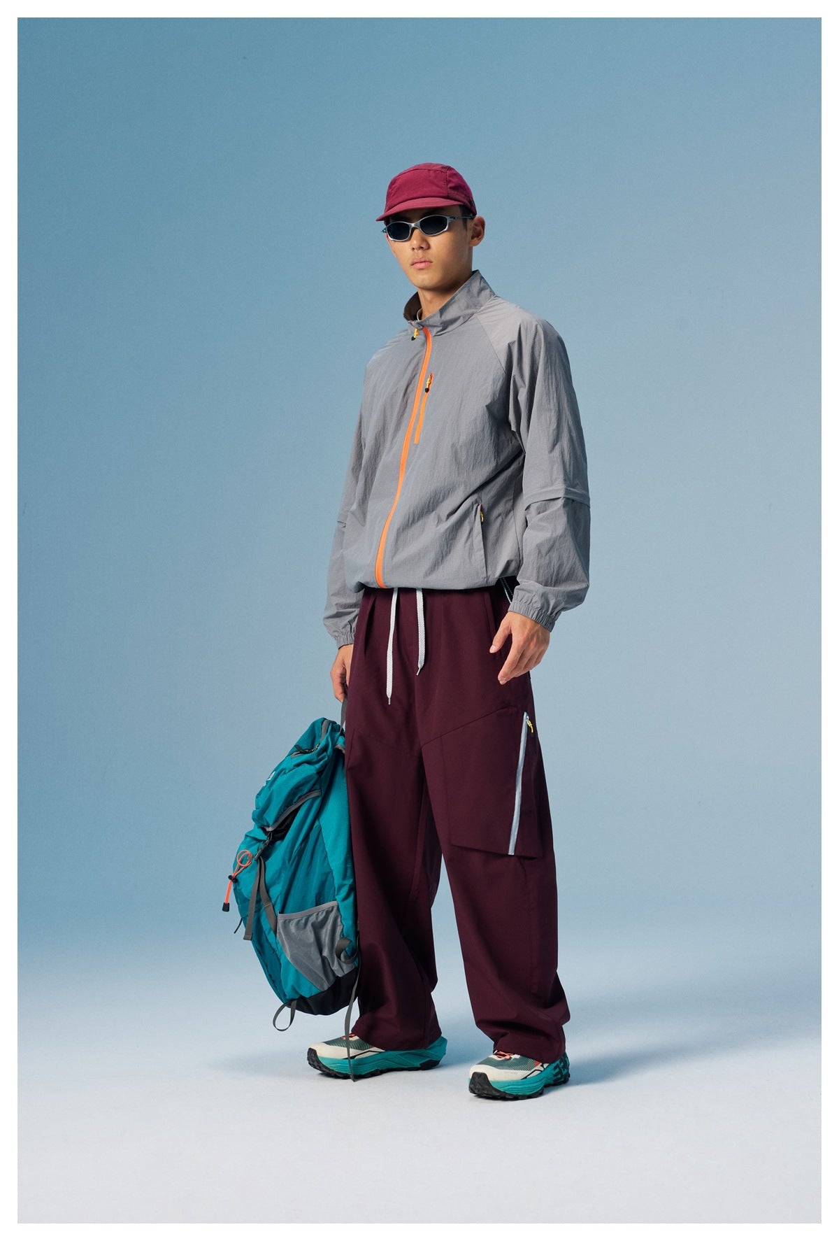 Full-length three-quarter view of a model in a gray orange-zip jacket and burgundy cargo pants, holding a teal backpack.
