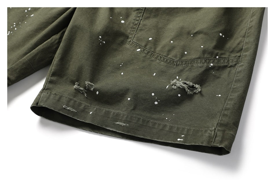 Close-up of olive cargo pants showing white paint splatters, distressed tears, stitching, and a hem.