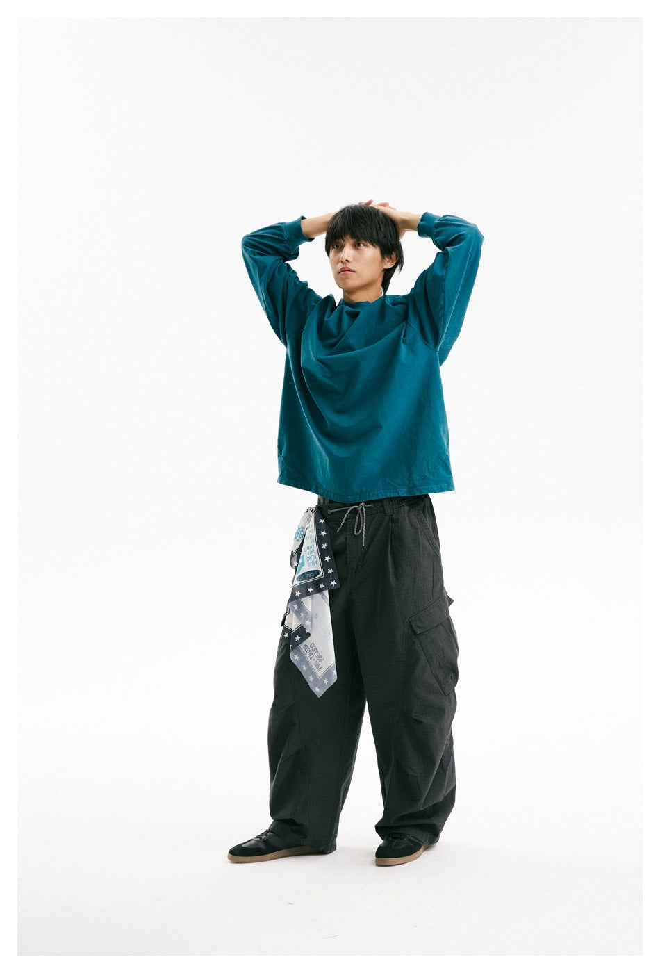 Model stretching with arms raised, wearing a teal raglan long-sleeve shirt and dark cargo trousers.