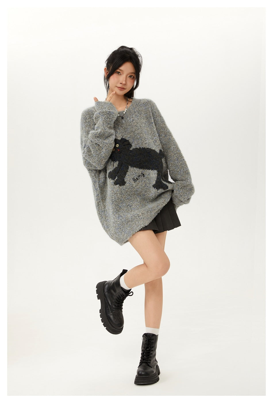 Model posing in a gray fuzzy knit sweater with a dark animal motif, black pleated skirt and lace-up boots