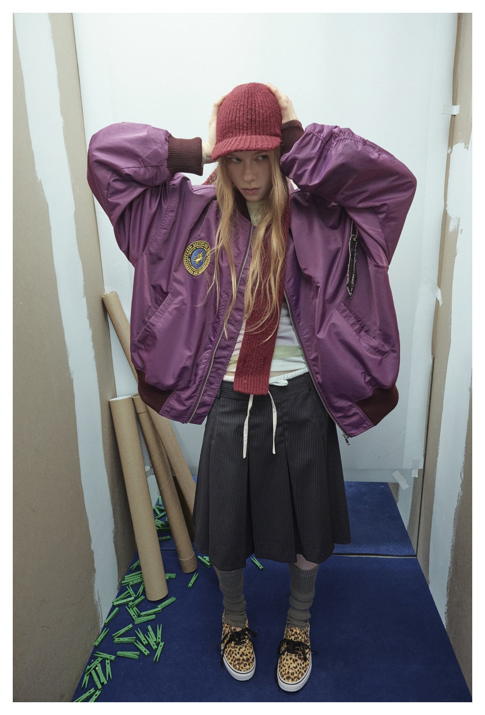 Model wearing an oversized purple bomber jacket with dark ribbed cuffs and hem, a round chest patch, and a red knit hat.