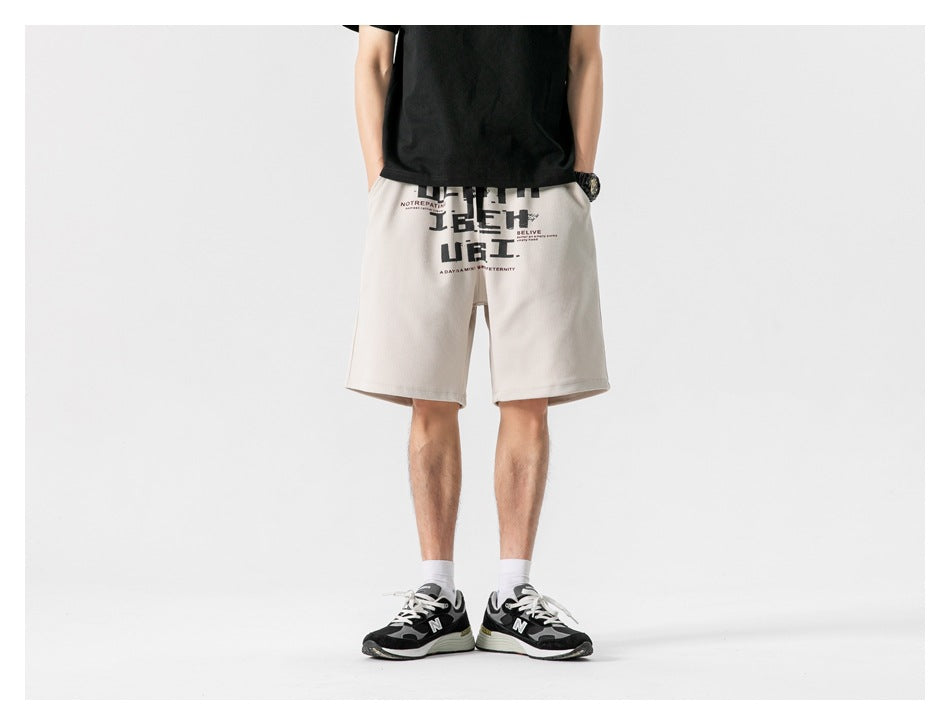 Model wearing khaki graphic-print drawstring shorts with a black tee and sneakers, front view, hands in pockets