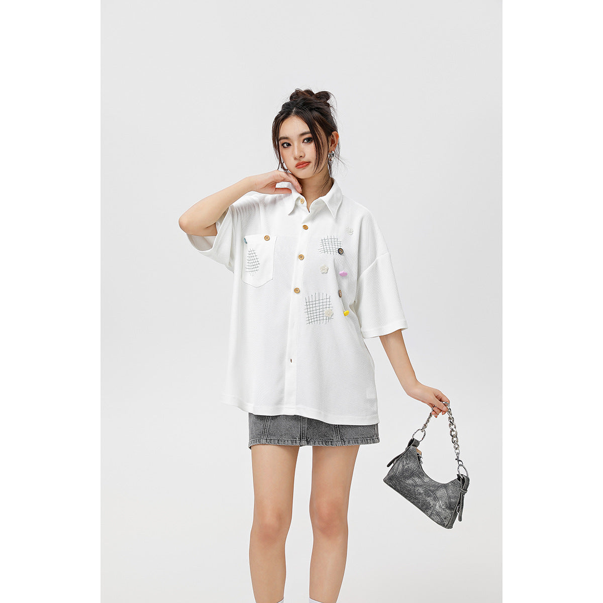Model wearing a white oversized short-sleeve button-up shirt with a chest pocket, grid patches and small colorful accents.