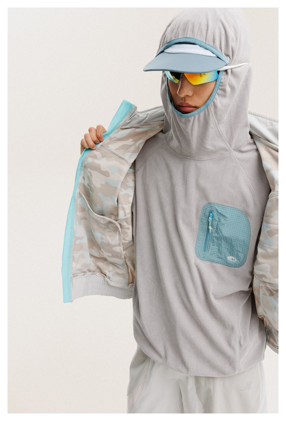 Close-up of a model opening a gray jacket to show its pale camouflage lining over a gray top with turquoise chest pocket.