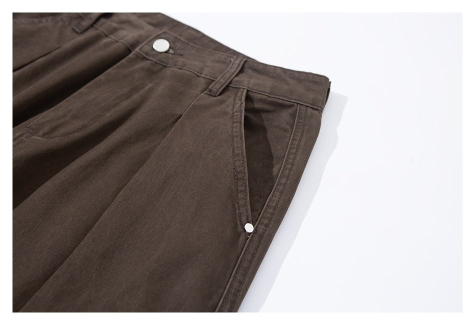 Angled detail of dark brown pants showing the waistband button, pleat and side pocket with a metal stud.