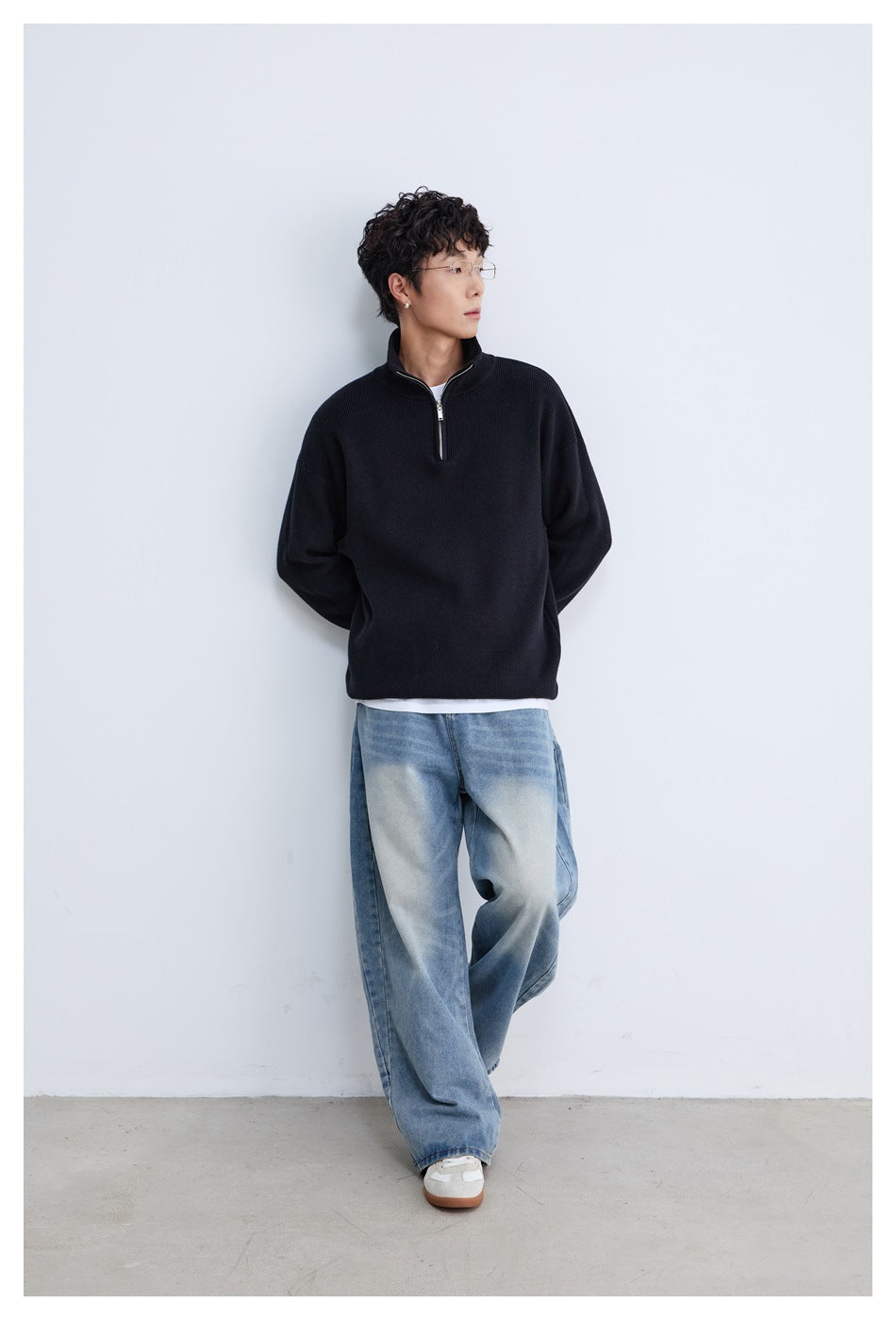 Model standing in a black quarter-zip sweater and loose blue jeans, looking to the side.