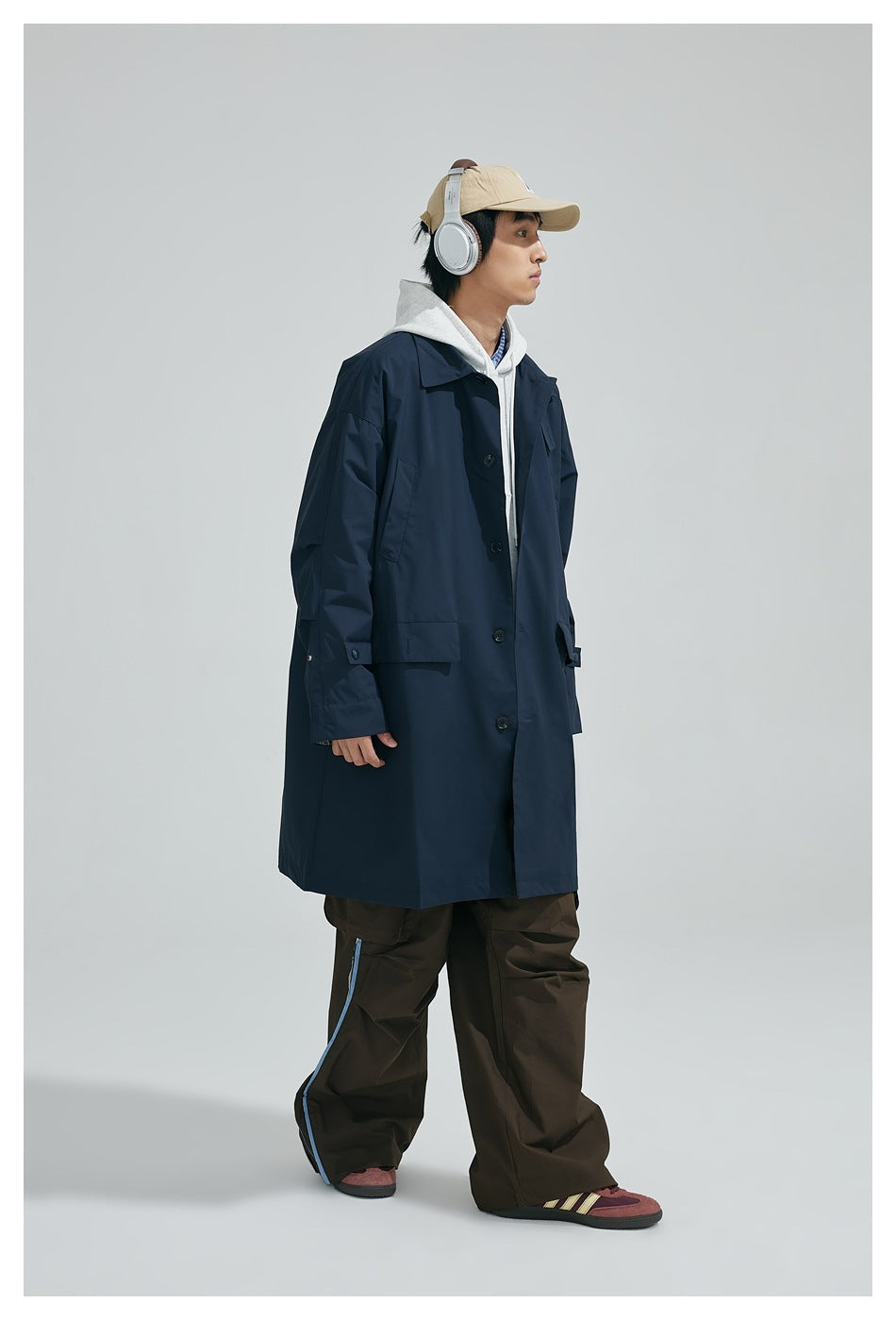 Side view of a model in a navy knee-length coat with flap pockets over a gray hoodie and brown wide-leg pants